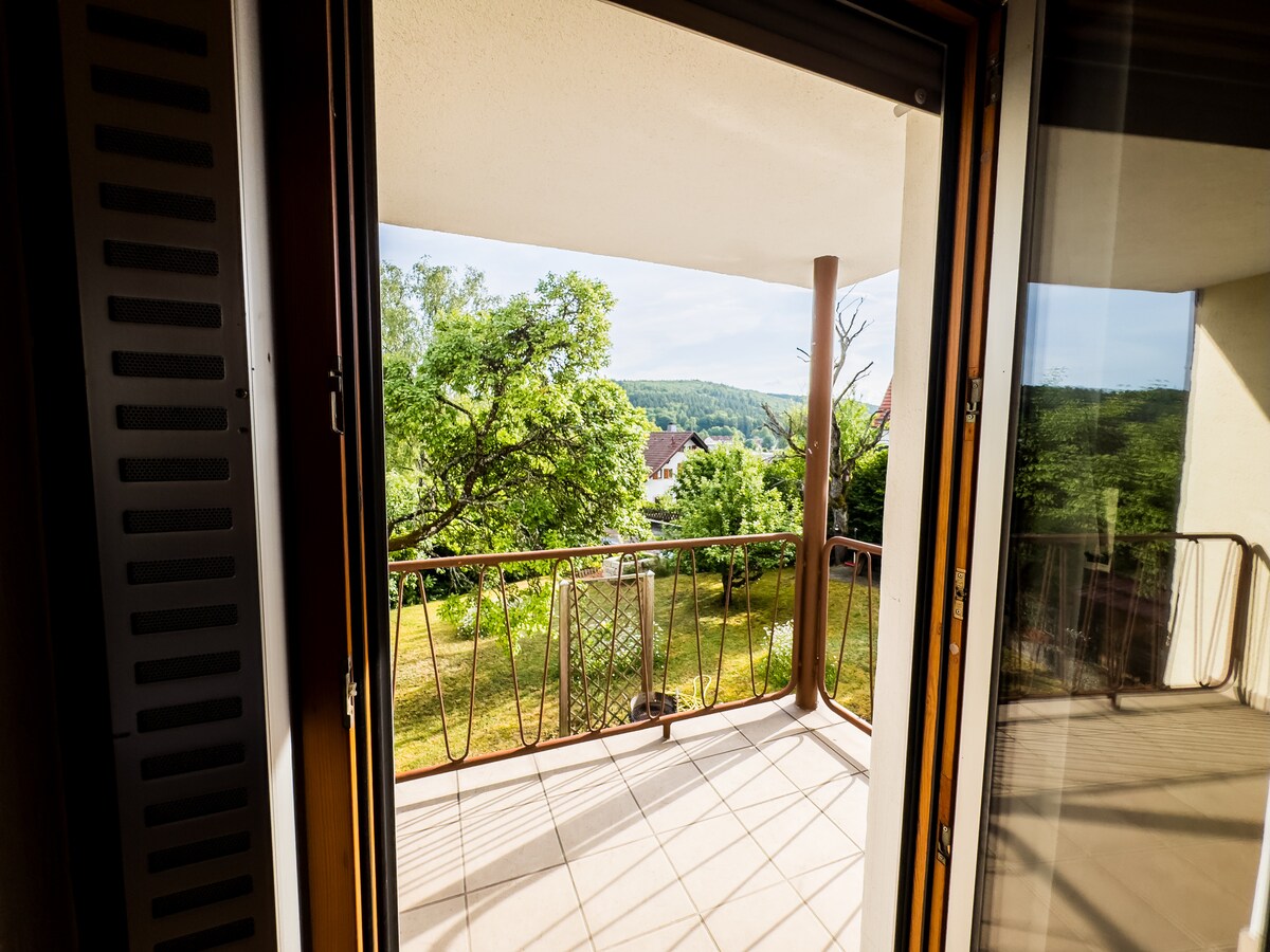 A view from the balcony is presented, showcasing a well-maintained garden with lush trees and a gentle slope in the background. The spacious balcony features a wrought iron railing, allowing for an enjoyable outdoor experience while overlooking the greenery.