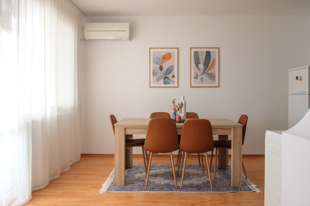 The dining area features a large wooden table surrounded by six upholstered chairs, placed on a softly patterned rug. Two framed botanical prints adorn the wall above the table, while natural light filters through sheer curtains, enhancing the inviting ambiance.