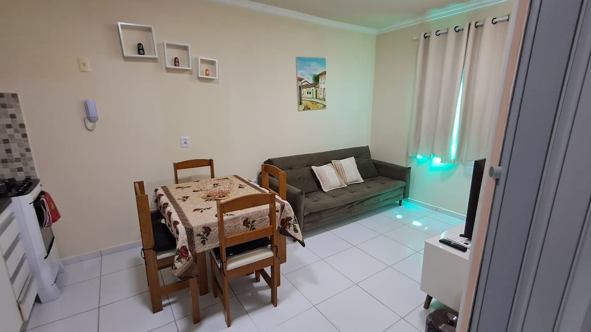 A comfortable living area features a light-colored sofa and a wooden dining table set for four. Shelves adorn the wall, displaying decorative items. A painting hangs to one side, and soft drapes frame the window, allowing natural light to brighten the space.
