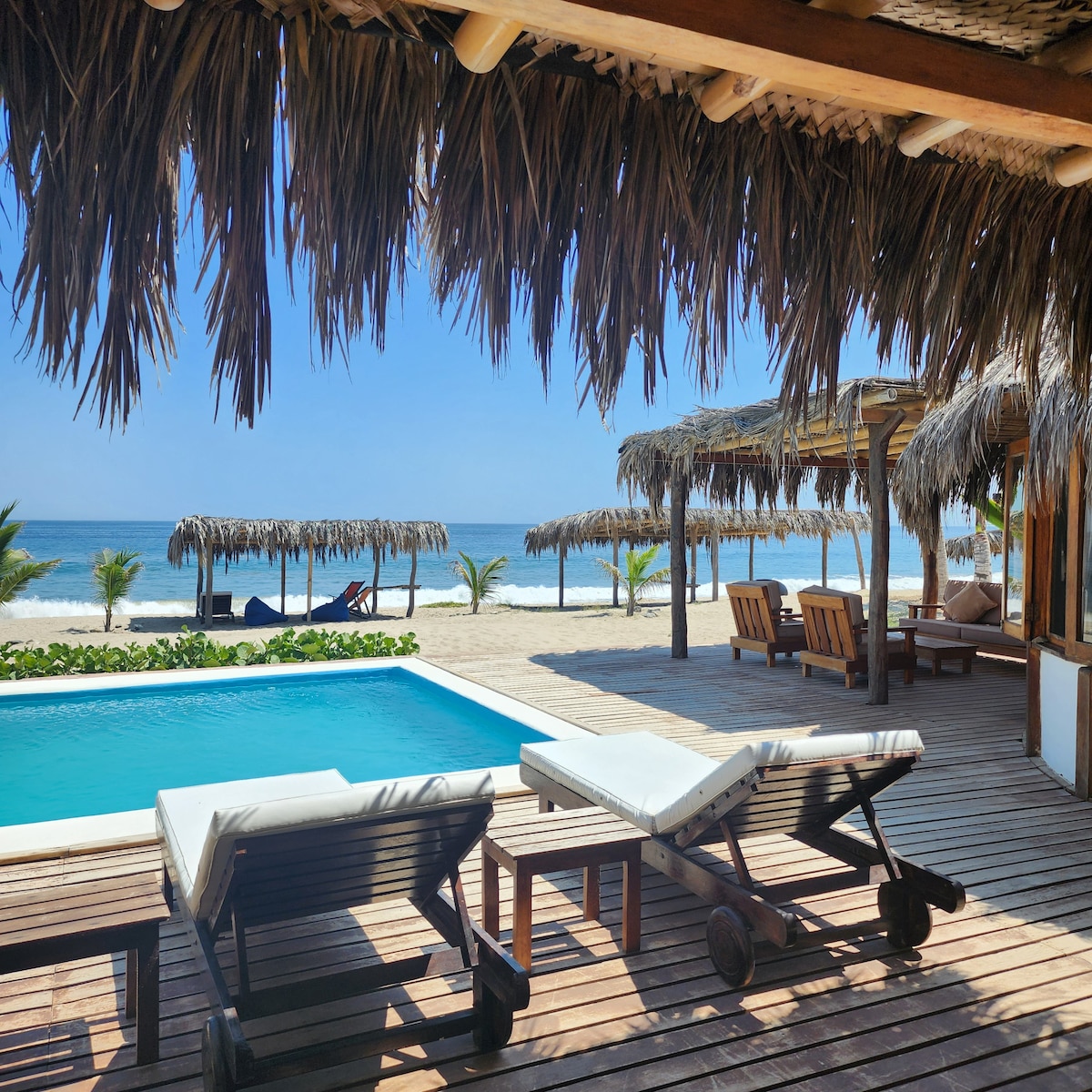 A serene pool area features white lounge chairs beside a turquoise pool. Thatched-roof structures provide shade along the beachfront, with palm trees harmoniously complementing the coastal setting. The calm sea stretches beyond, enhancing the tranquil atmosphere.