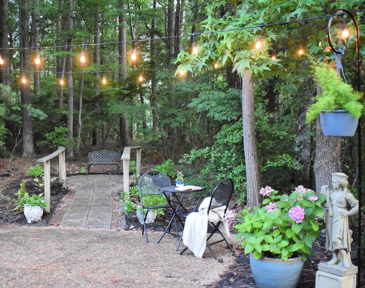 A charming outdoor patio area is depicted, featuring a small round table with two black chairs. String lights hang overhead, casting a warm glow. A pathway made of stone leads through lush greenery, accented by decorative pots with blooming flowers.