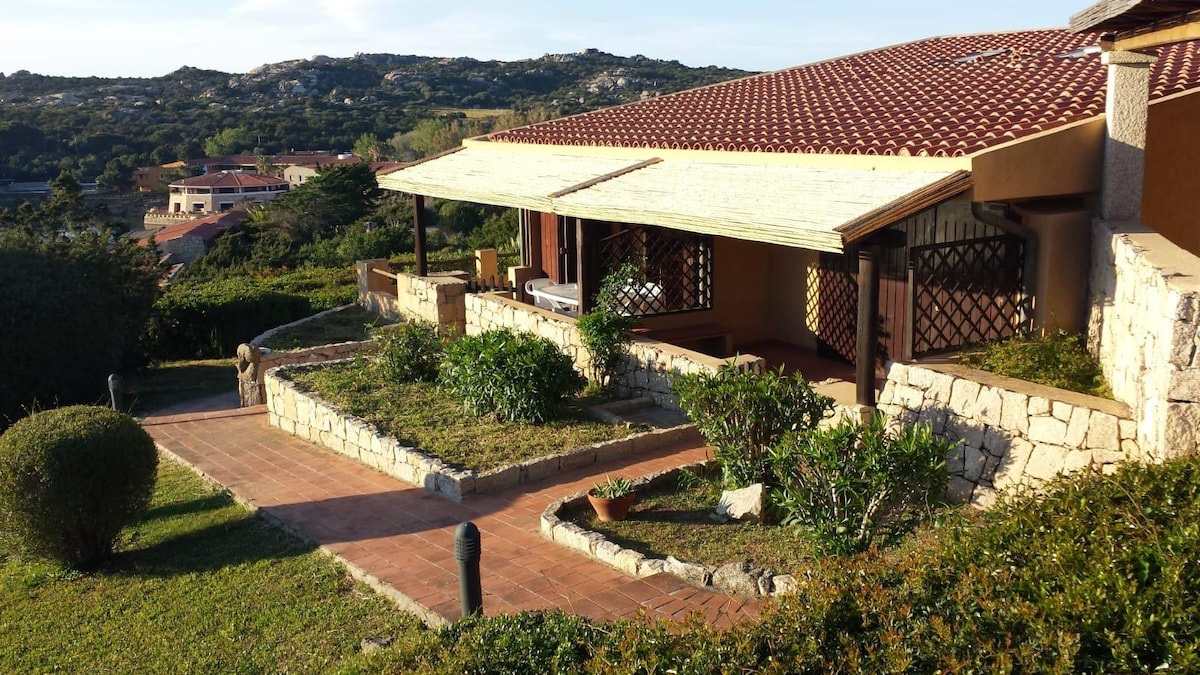 The exterior of the villa features a spacious veranda with a wooden roof. Lush greenery frames the property, with stone pathways leading to the entrance. Surrounding hills and a clear sky create a serene backdrop, evoking a sense of tranquility.