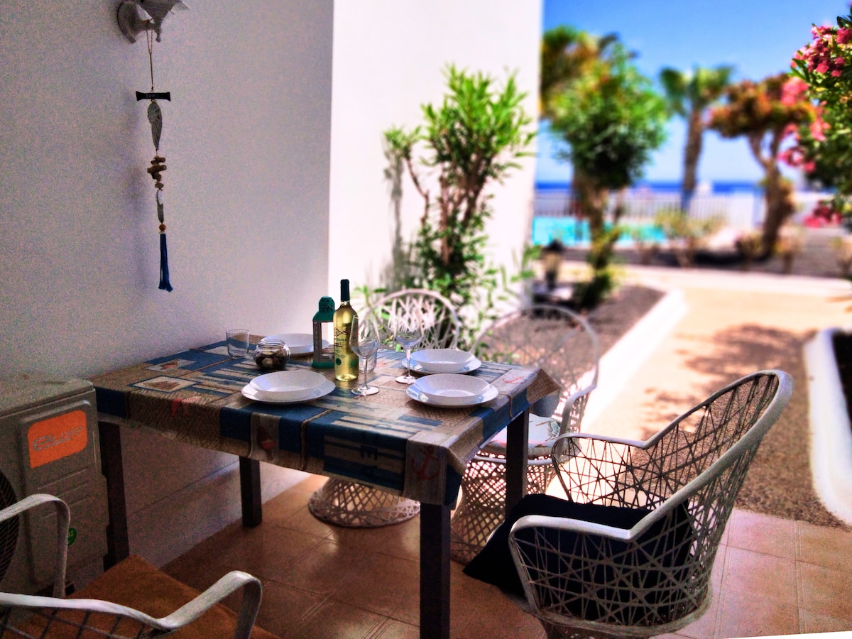 An outdoor dining area is presented with a rectangular table set for four. Two wine bottles and glasses are arranged, complemented by island views and lush greenery. The table is covered by a patterned tablecloth, inviting guests to enjoy meals in a serene atmosphere.