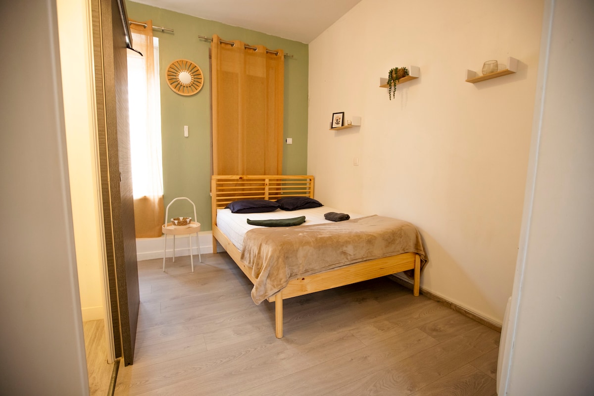 A bright sleeping area is furnished with a spacious wooden bed, covered with a soft beige blanket. Two textured pillows are neatly arranged on top. Natural light filters through sheer curtains, while a small bedside table holds a decorative item. Green accent walls provide a calming backdrop.