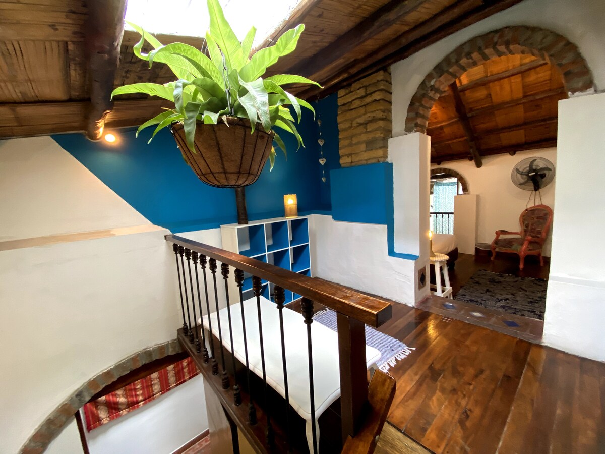 A bright, airy landing overlooks a spacious area below, adorned with a large potted plant hanging from the wooden ceiling. The white walls are complemented by blue accents, and light from a nearby window illuminates the warm wooden flooring.