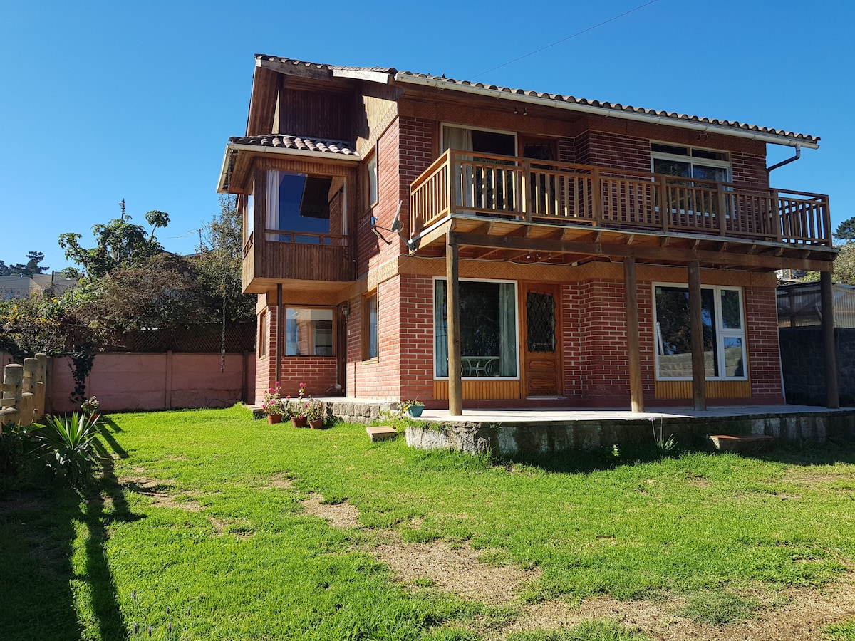 The two-story house features a brick facade with wooden accents and a spacious lawn. Large windows allow natural light to fill the interior. A balcony with wooden railings overlooks the garden, enhancing the outdoor living space.