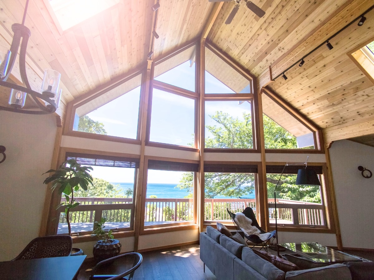 The spacious living area features large triangular windows that provide a panoramic view of the surrounding nature and sea. Natural wood beams accentuate the ceiling, while sunlight streams through the windows, illuminating the room. A comfortable sofa is arranged to enjoy the view.