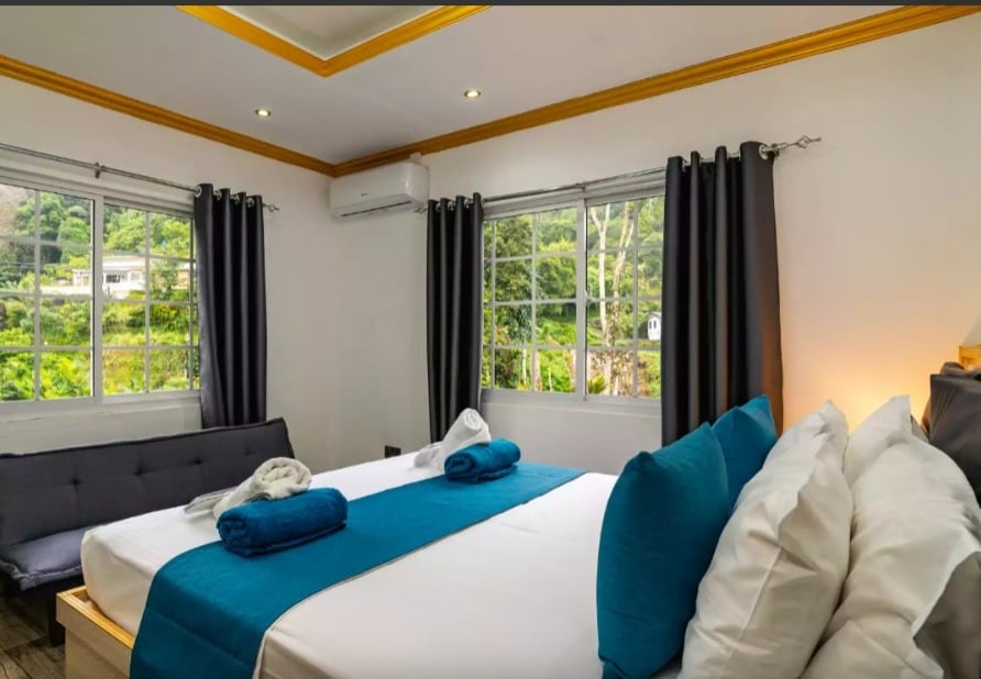 A bright bedroom features large windows allowing natural light and views of the surrounding greenery. A spacious bed is adorned with neatly arranged pillows and a vibrant blue blanket. A comfortable seating area with a gray sofa adds to the inviting ambiance.