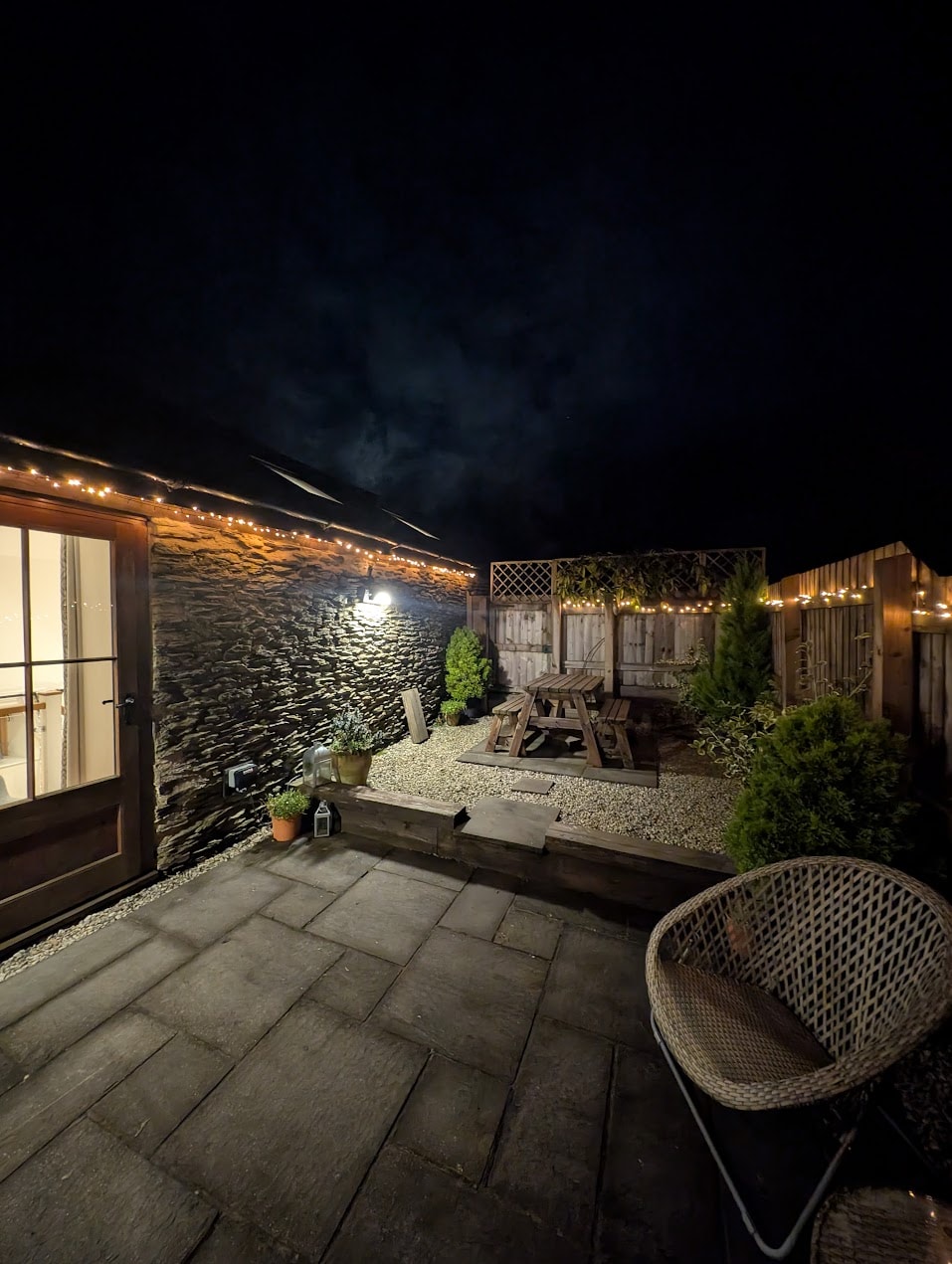 A private outdoor terrace is illuminated by soft hanging lights, creating a serene atmosphere. A wooden picnic table is surrounded by greenery and gravel pathways. Nearby, a cozy chair adds to the seating options, complementing the stone wall backdrop.