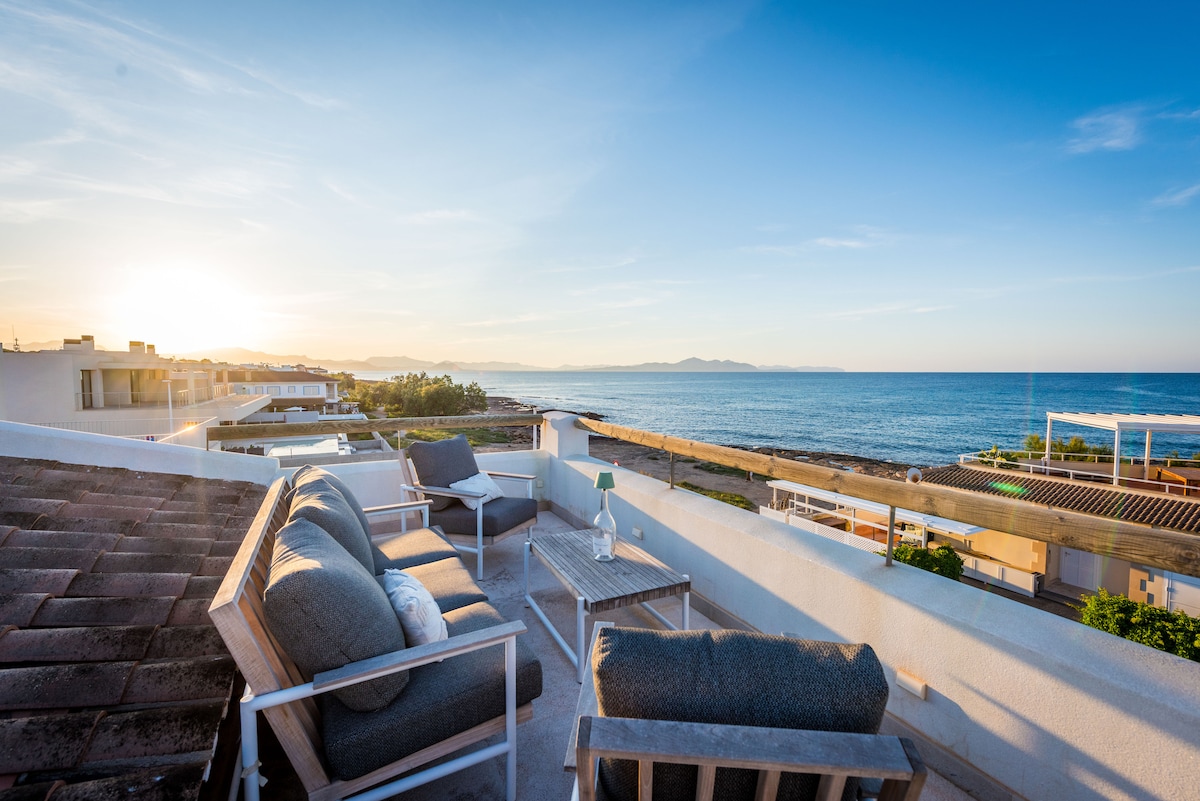 A private rooftop terrace is showcased with comfortable lounge seating overlooking the sea. The landscape features a clear horizon, with the sun setting in the distance. The serene ocean can be seen, enhancing the tranquil atmosphere of the outdoor space.