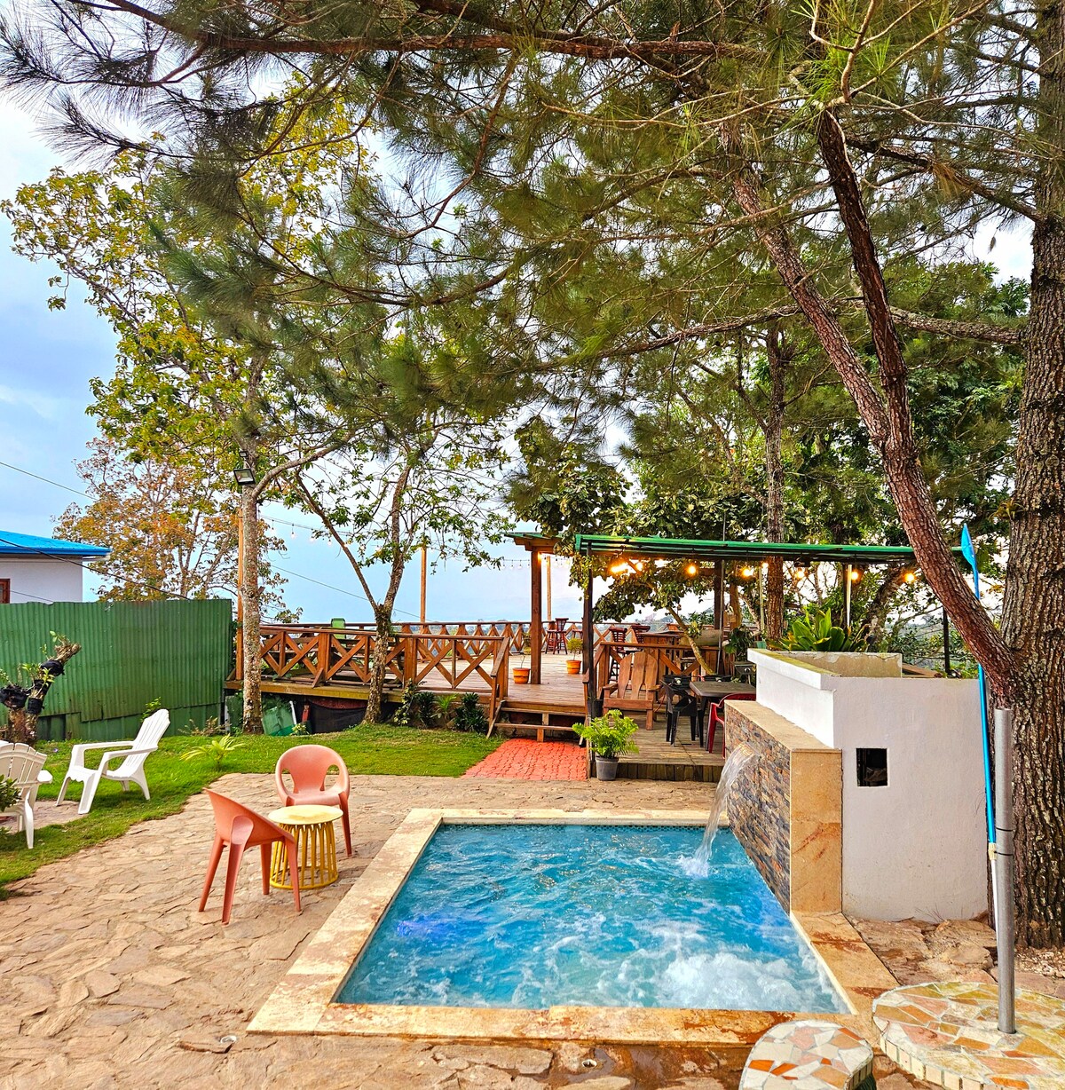 A private outdoor area features a small pool with a waterfall feature, surrounded by trees. Colorful chairs are placed near the pool, and a wooden deck is visible in the background, providing an inviting space for relaxation. The landscaped garden enhances the natural setting.