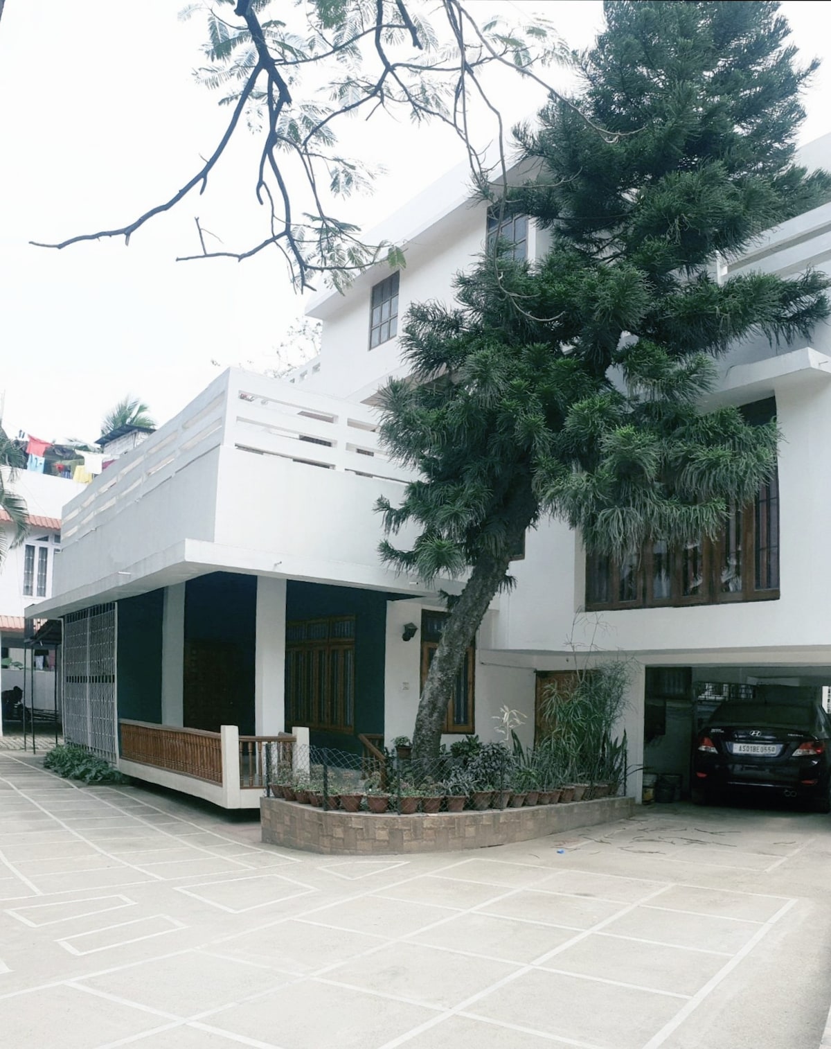 The exterior features a modern, two-story structure with a spacious overhang and a large tree providing shade. A parked vehicle is visible in the designated parking area, with a clean driveway and a landscaped border that includes plants and greenery.