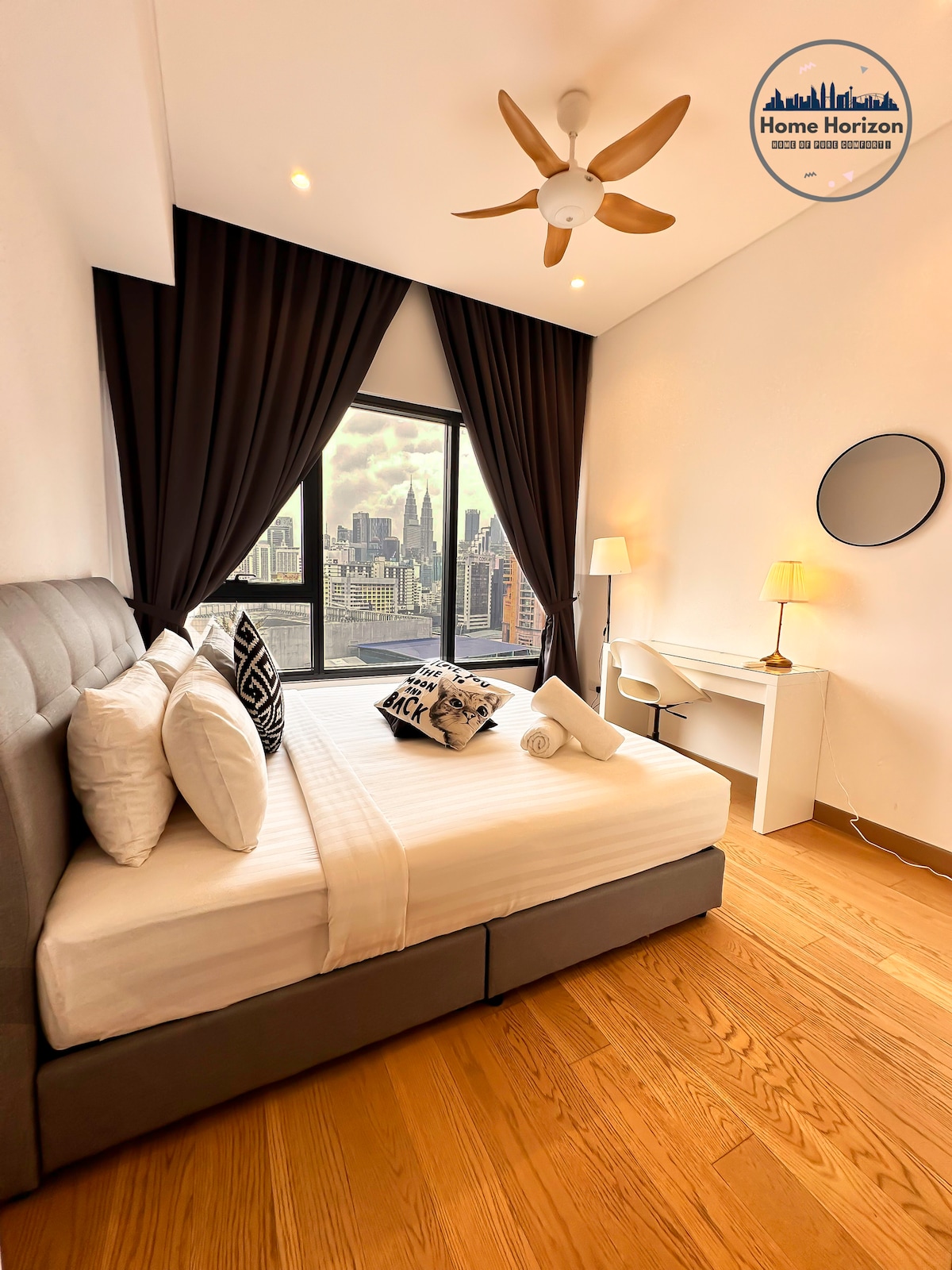 A cozy bedroom features a spacious bed with crisp white linens, accompanied by decorative pillows. Large windows offer an expansive view of the city skyline, while a modern desk and chair provide a functional workspace. Soft curtains frame the windows and a stylish ceiling fan adds to the ambiance.