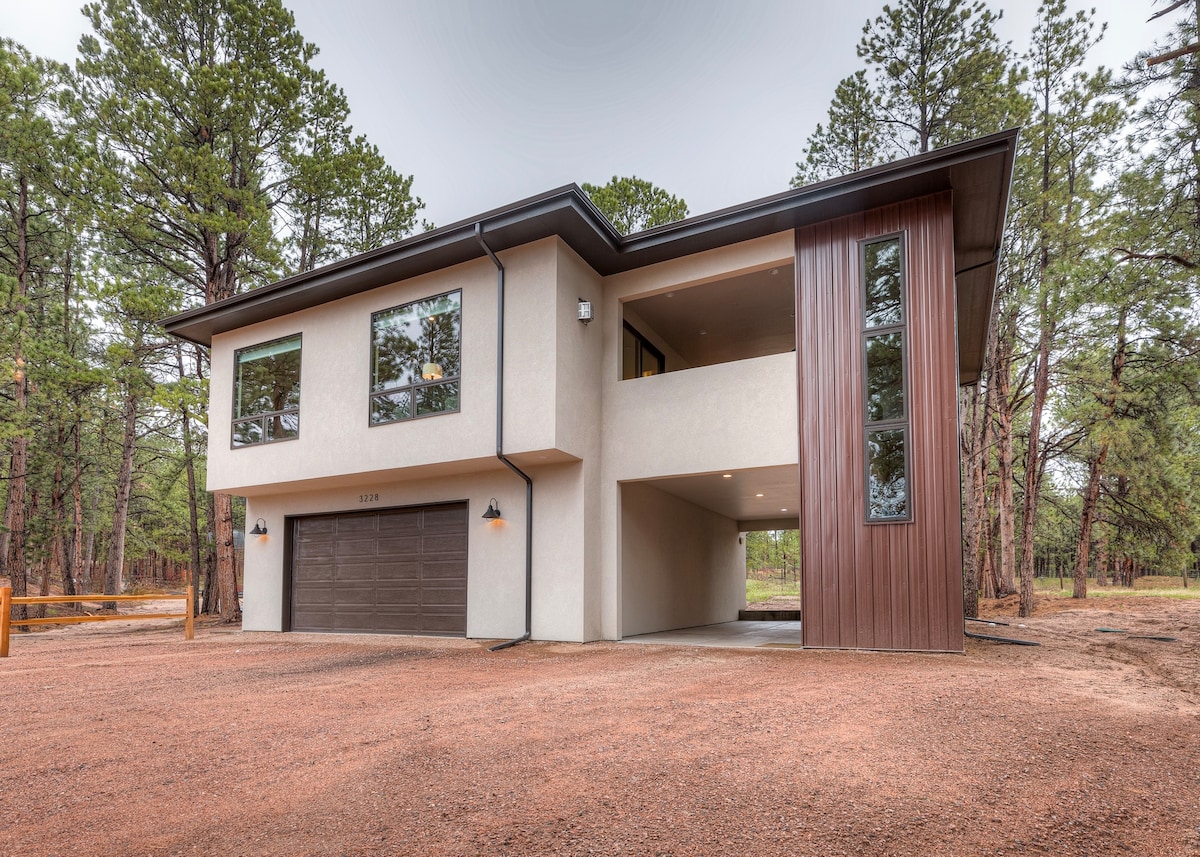 The exterior of a modern two-story guest house is shown, featuring large windows and a combination of light and dark materials. The structure is situated amidst tall pine trees on a gravel driveway, with a single carport located beneath the upper level.