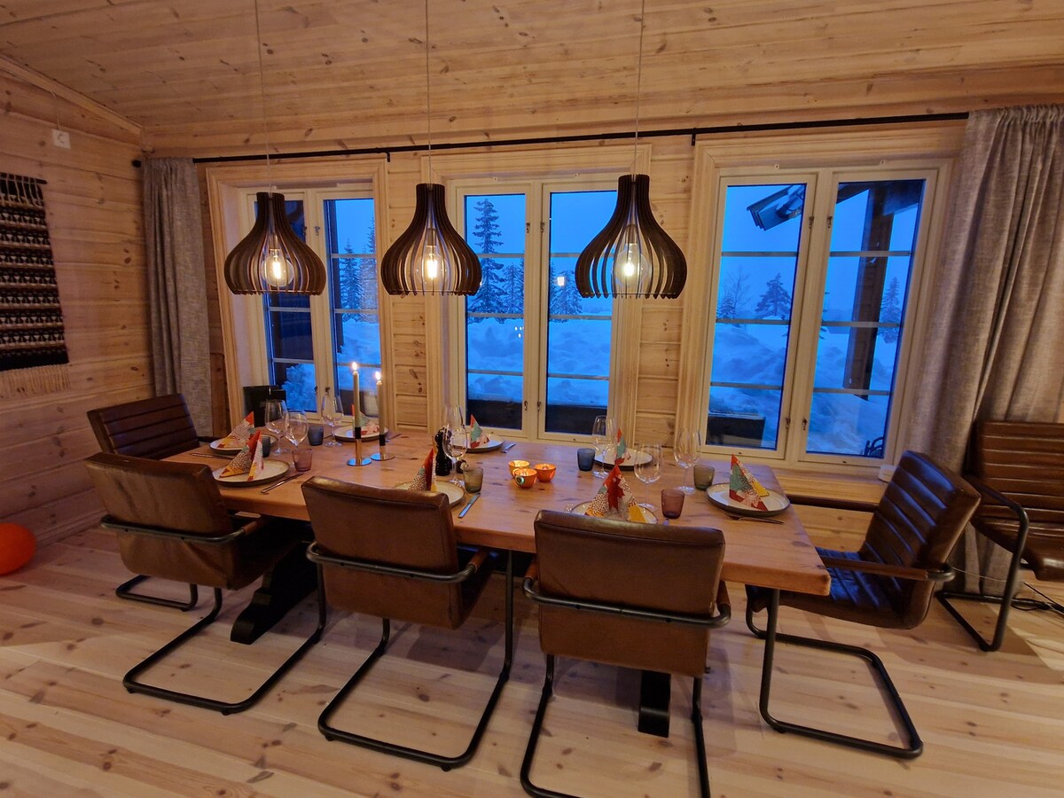 A dining area features a large wooden table surrounded by modern brown chairs. Soft candlelight is present on the table, with plates set for a meal. Large windows allow for views of the snowy outdoors, complemented by stylish hanging light fixtures.