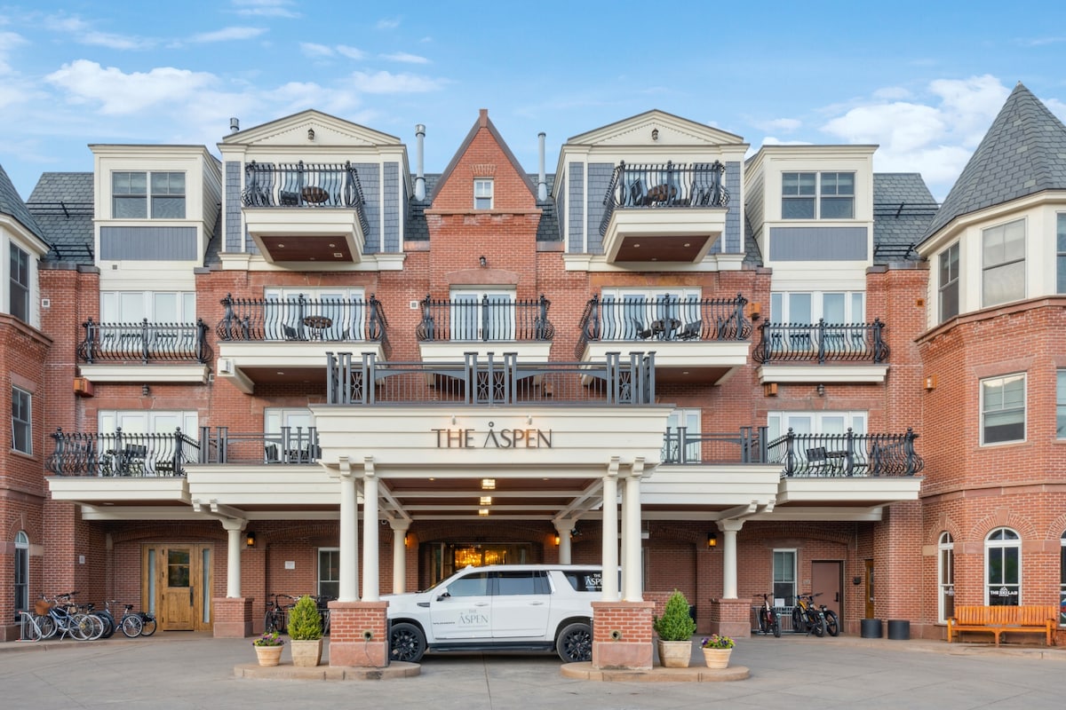The exterior of The Aspen Mountain Residences is captured, showcasing a charming brick facade with balconies. The entrance is marked by a welcoming portico, and bicycles are parked neatly in front. A white vehicle is positioned just off the main entrance.