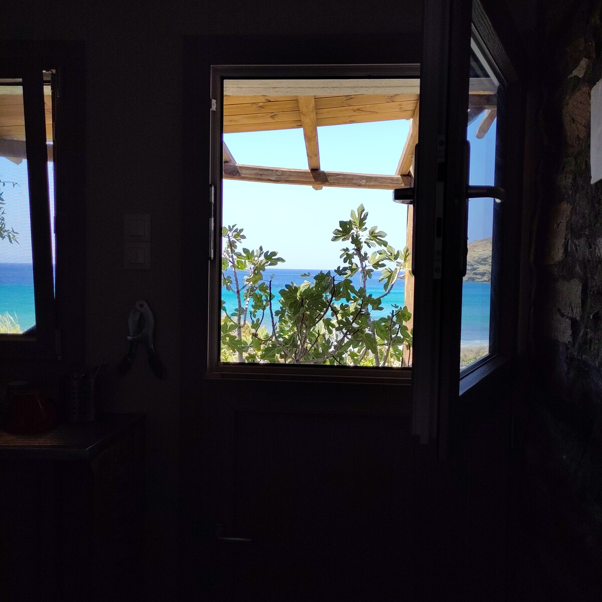 A window frames a view of the tranquil sea, with a lush fig tree partially visible outside. Sunlight pours in, highlighting the natural stone walls and wooden accents of the bungalow, creating an inviting atmosphere that connects indoors with the shore.