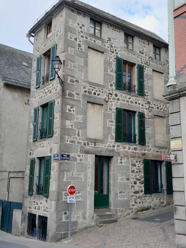 The exterior of a historic three-story stone building is depicted, featuring green shutters and decorative wrought iron balconies. A street sign is visible, along with a 'Stop' sign. The structure reflects local architectural style and is situated in a city center environment.