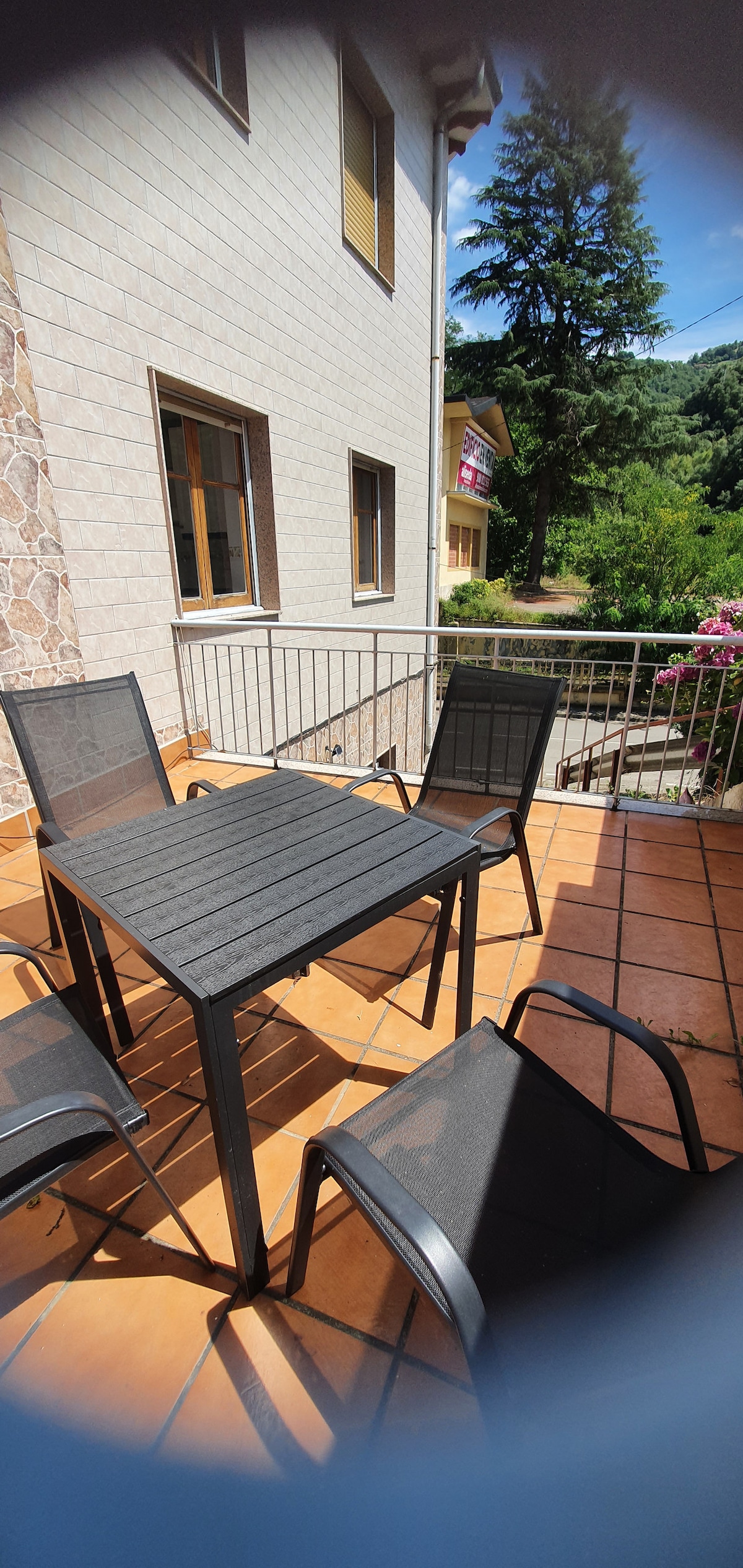 A terrace featuring a square table surrounded by several chairs is visible. The flooring consists of terracotta tiles, and a railing provides safety. Lush greenery and flowers can be seen in the background, contributing to a tranquil outdoor setting.
