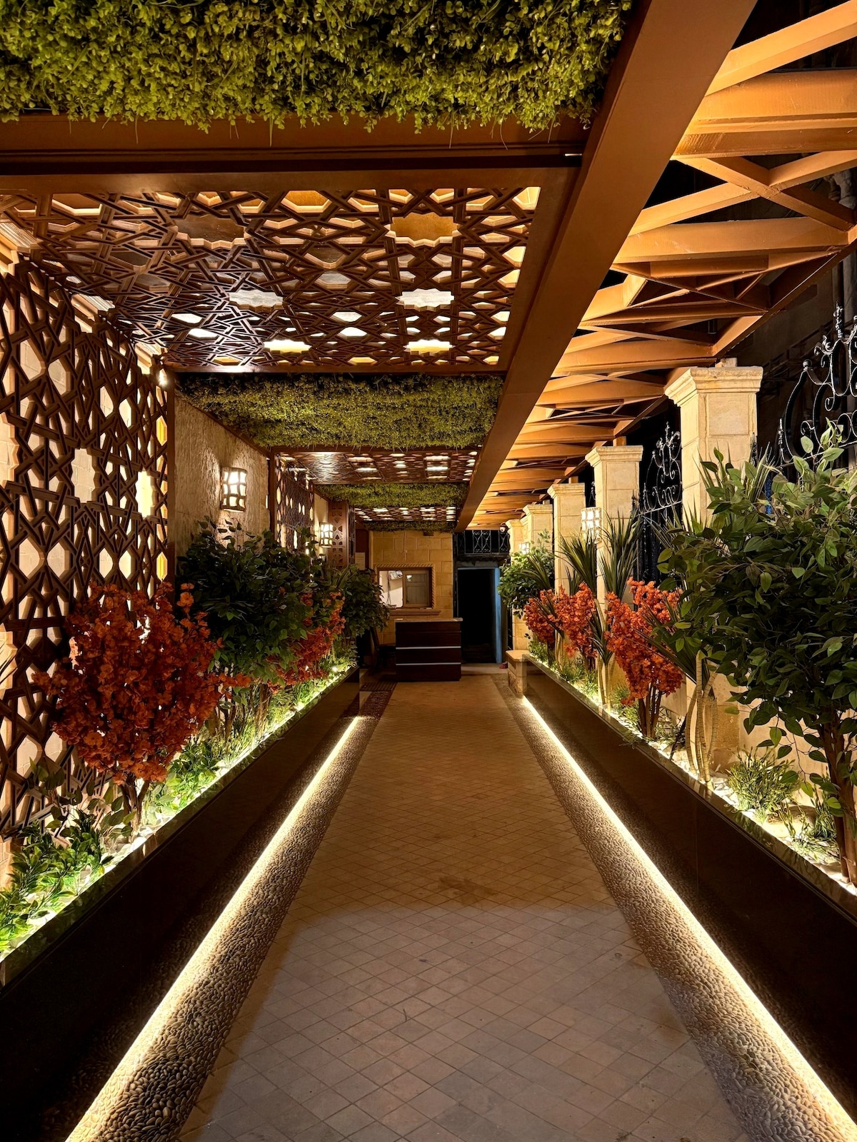 An elegant corridor is lined with decorative latticework and lush greenery, providing a serene atmosphere. Soft, warm lighting highlights the path, and vibrant plants in planters add color to the space, creating a welcoming and tranquil entryway.