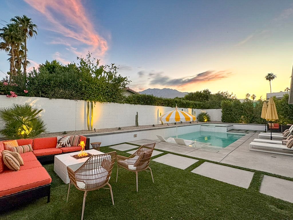 An outdoor space features a custom-designed pool with a tanning ledge, surrounded by lounge chairs and patio furniture. Lush greenery and palm trees provide a backdrop, with vibrant sunset hues lighting the scene. A shaded area includes an umbrella, while seating arrangements invite relaxation.