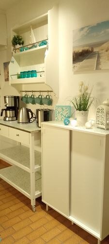 A well-organized kitchenette area is presented, featuring a coffee maker, kettle, and multiple containers. Shelves display turquoise glassware and decorative items, accompanied by a potted plant. A panoramic beach photo adds a serene touch to the space.