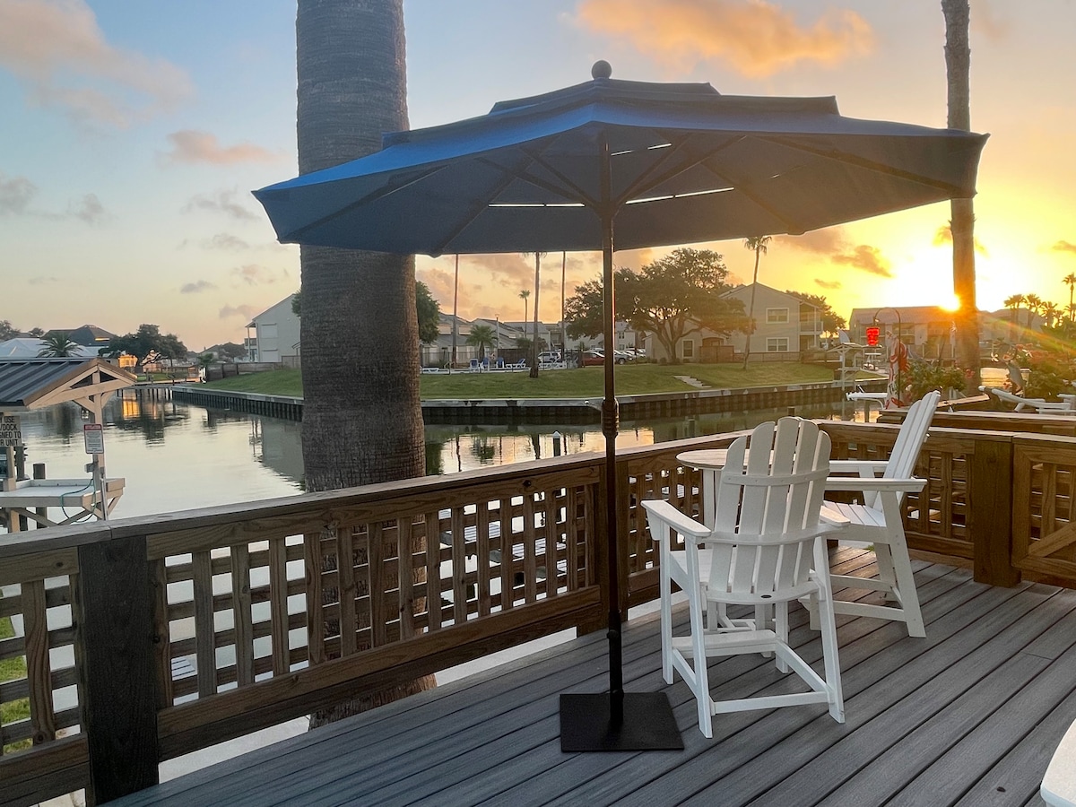 A serene patio scene features a blue umbrella providing shade over two classic white Adirondack chairs, positioned on a wooden deck. A tranquil waterway is visible in the background, reflecting the warm hues of a sunset, with palm trees framing the view.