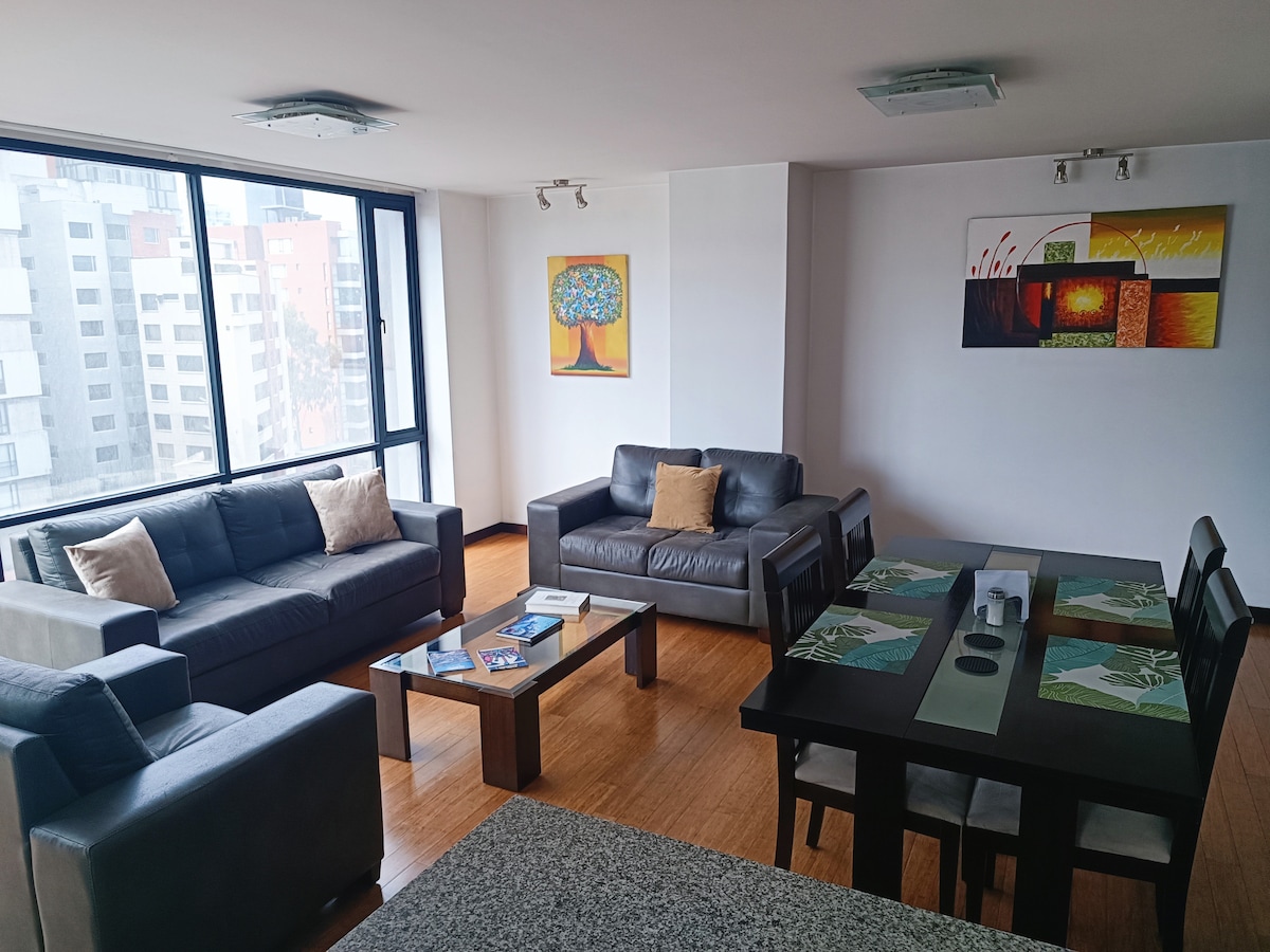 A spacious living area features two comfortable sofas, accompanied by a coffee table, and a dining table set for six. Large windows provide ample natural light and city views. Colorful artwork adorns the walls, adding a touch of character to the space.