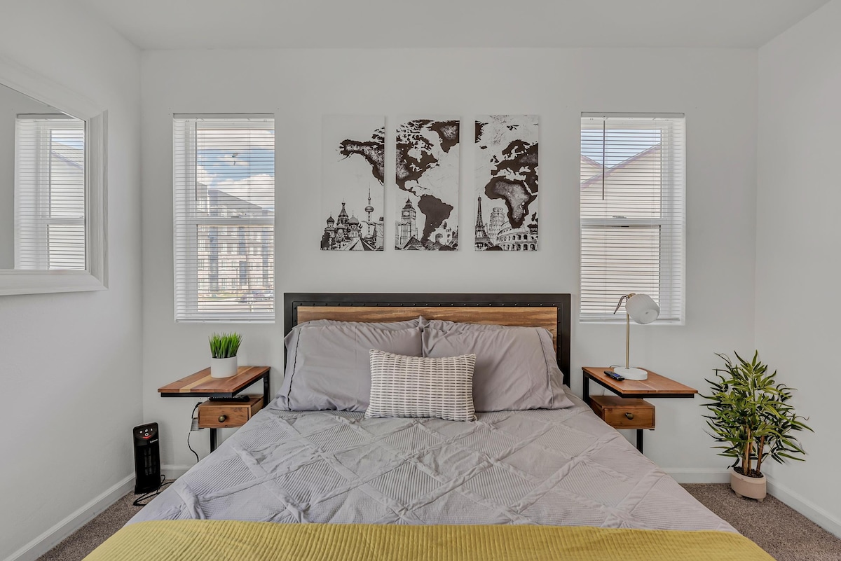 The bedroom features a queen-sized bed with gray bedding and a decorative yellow throw blanket. Two bedside tables are positioned on each side, supporting bedside lamps. World map artwork adorns the wall above the bed, while three windows provide natural light, adorned with blinds.