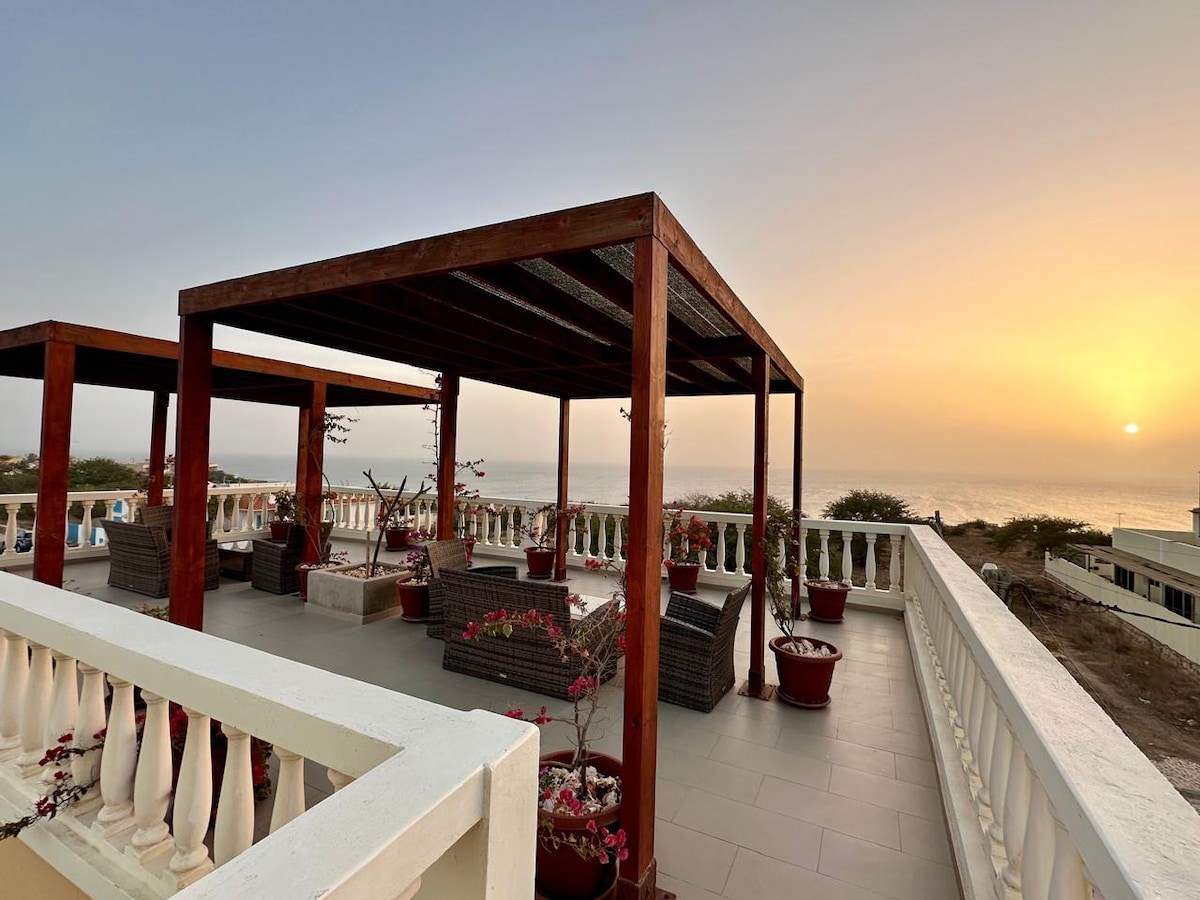 An open terrace with wooden pergolas provides a scenic view of the ocean and a vibrant sunset. Comfortable seating is arranged beneath the structure, surrounded by blossoming plants in pots, enhancing the outdoor space.