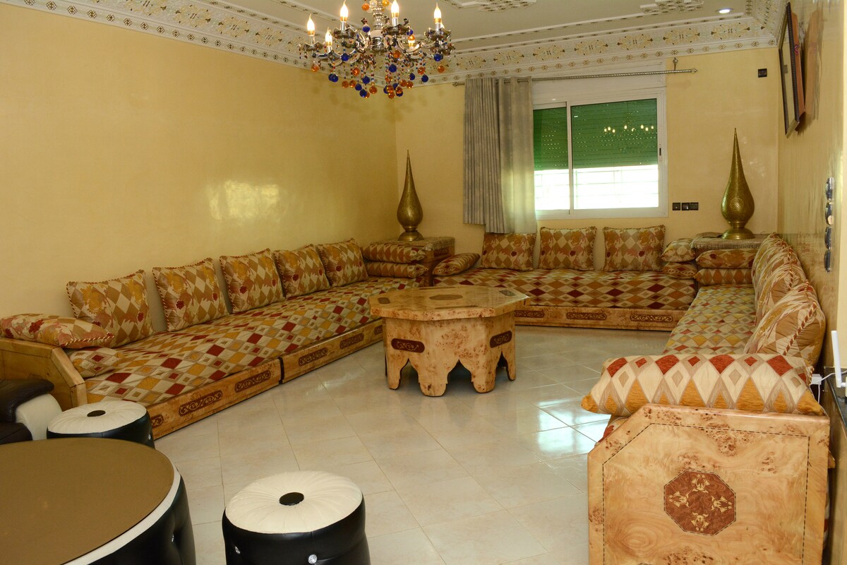 A spacious living area features a large, intricately designed seating surrounded by decorative cushions. A central table complements the seating, and a colorful chandelier hangs from the ceiling. Natural light enters through a window draped with sheer curtains.