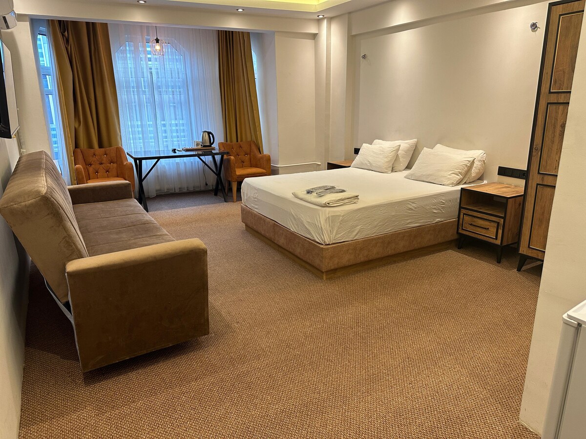 A spacious room features a large double bed and a comfortable seating area with a sofa and armchairs. A desk is positioned next to the window, which is dressed with light curtains. Illuminated by overhead lighting, the room offers a welcoming and bright ambiance.