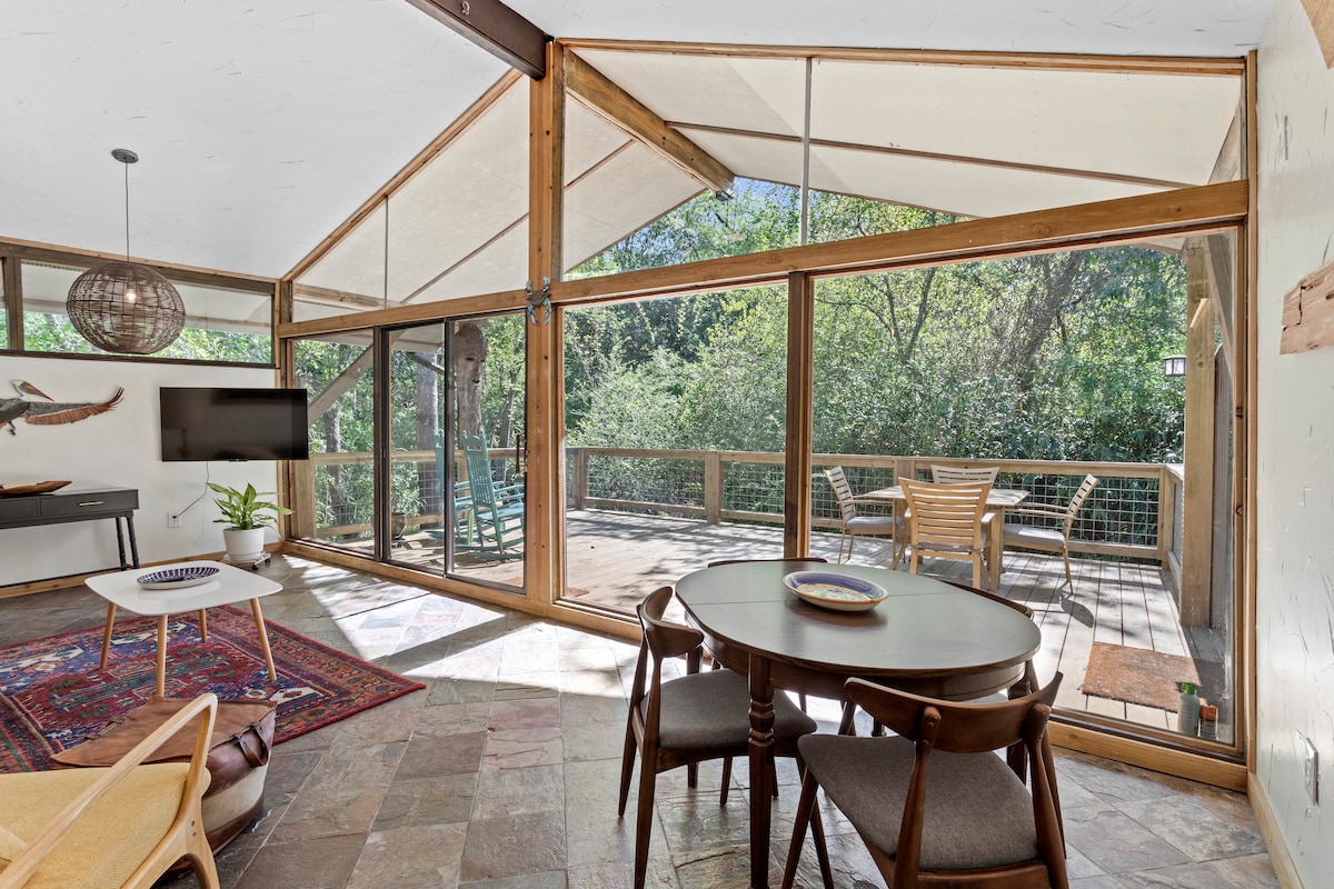 A bright living area is framed by floor-to-ceiling windows, inviting natural light and showcasing views of the greenery outside. A round dining table with wooden chairs sits comfortably in the space. A private deck is visible beyond, providing an outdoor seating area.