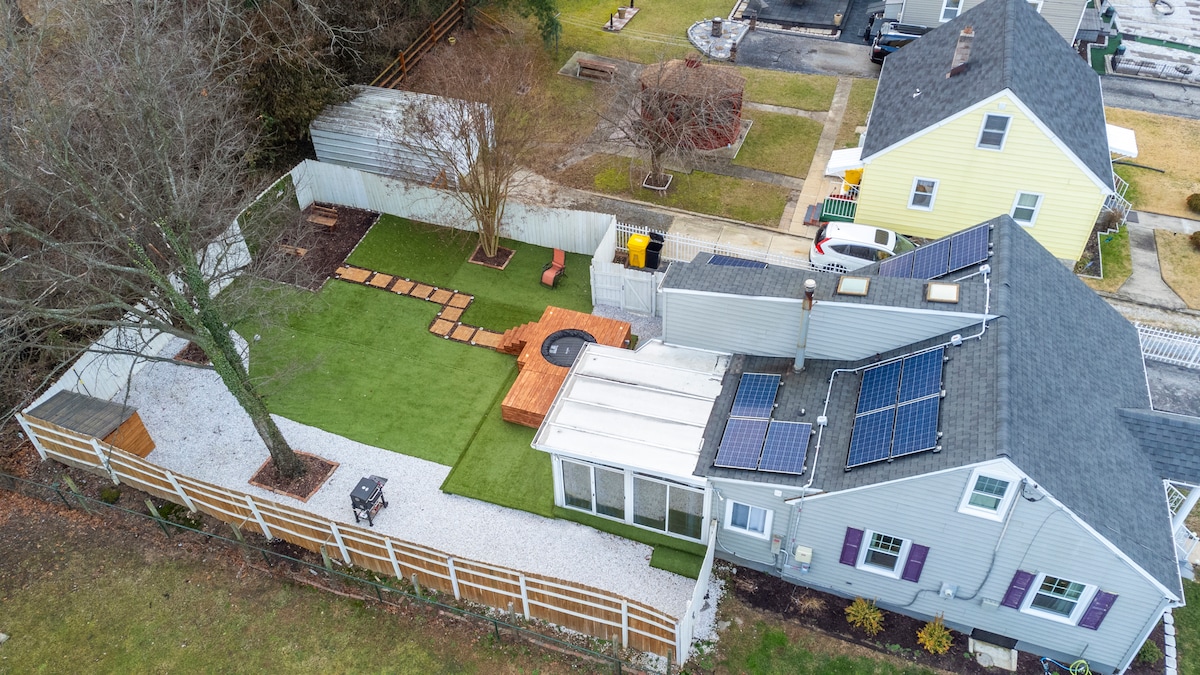 An aerial view captures a spacious outdoor area featuring a well-maintained green lawn, wooden deck, and hot tub. A gravel path leads through the yard, bordered by trees and fenced areas. Solar panels are visible on the rooftops of the home.