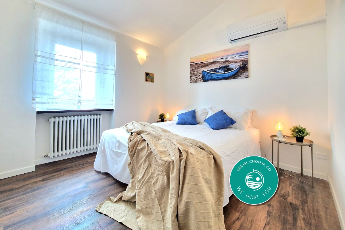 A bright sleeping area is presented with soft white bedding and two blue accent pillows. Natural light fills the room through a window, complemented by warm lighting from bedside lamps. A piece of artwork depicting a boat hangs above the bed, enhancing the calming ambiance.