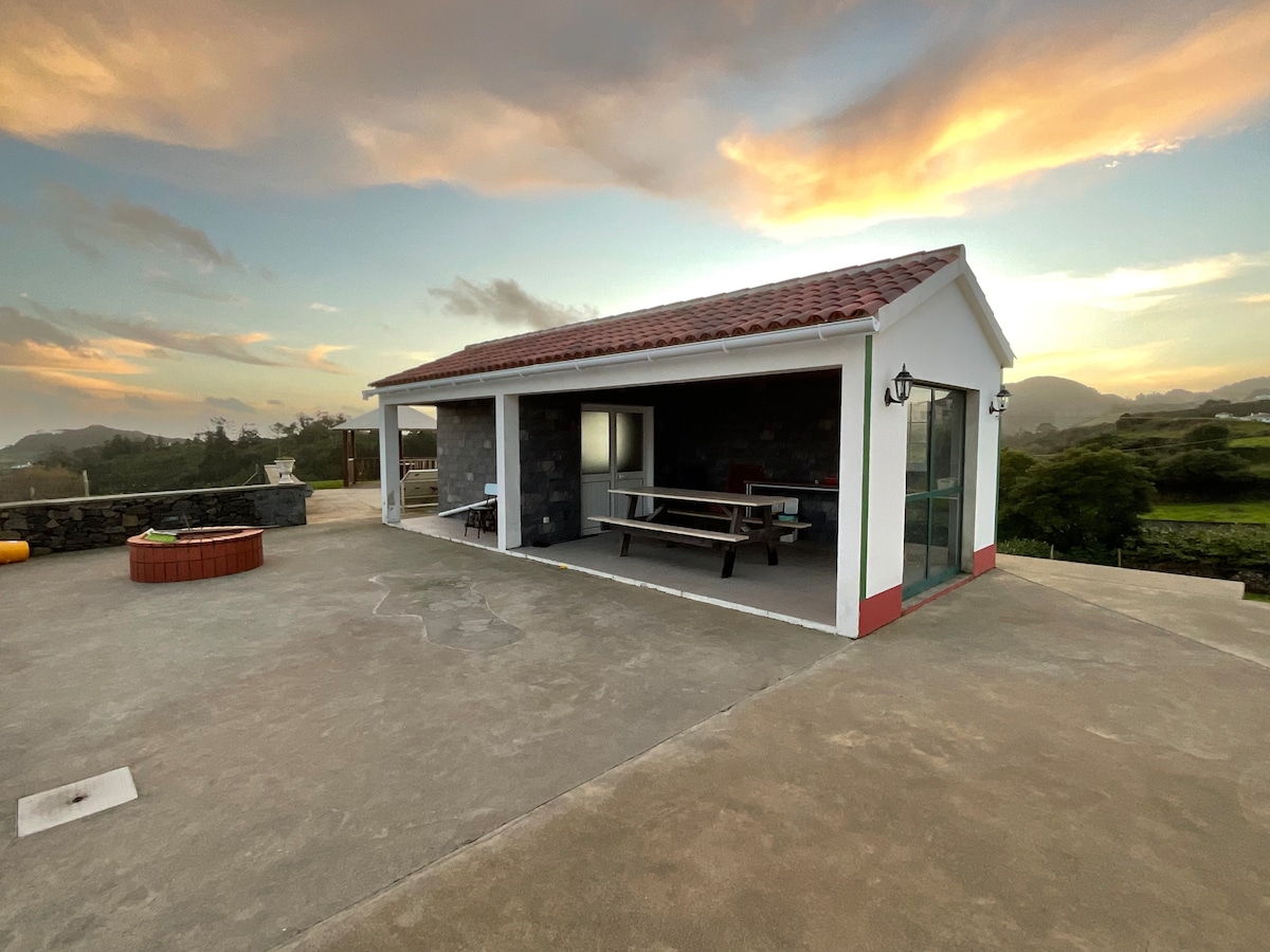 An outdoor view of a contemporary building with a tiled roof and stone accents, featuring large glass doors. A picnic table is positioned on a concrete patio, with a scenic landscape visible in the background, illuminated by a warm sunset.