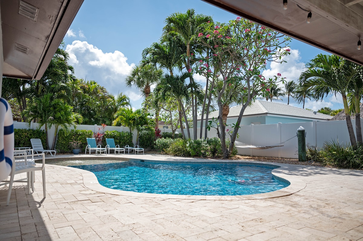 A tranquil backyard oasis features a sparkling saltwater pool surrounded by a stone deck. Lush tropical trees and vibrant pink flowers provide a serene backdrop, with several lounge chairs arranged nearby for relaxation.