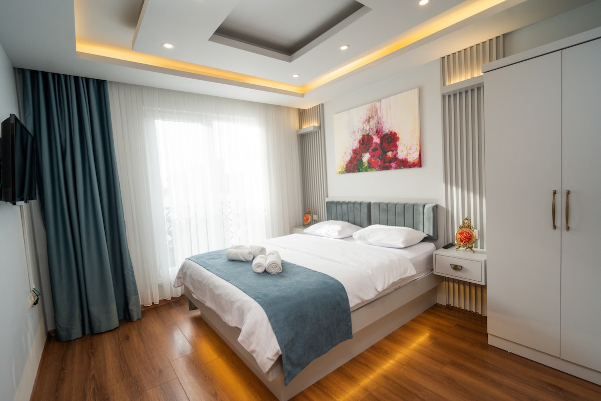 A well-appointed bedroom features a spacious bed with neatly arranged linens and two rolled towels on top. A calming color palette is created with grey accents and soft lighting. Large windows allow natural light, complemented by sheer curtains and decorative wall art above the headboard.