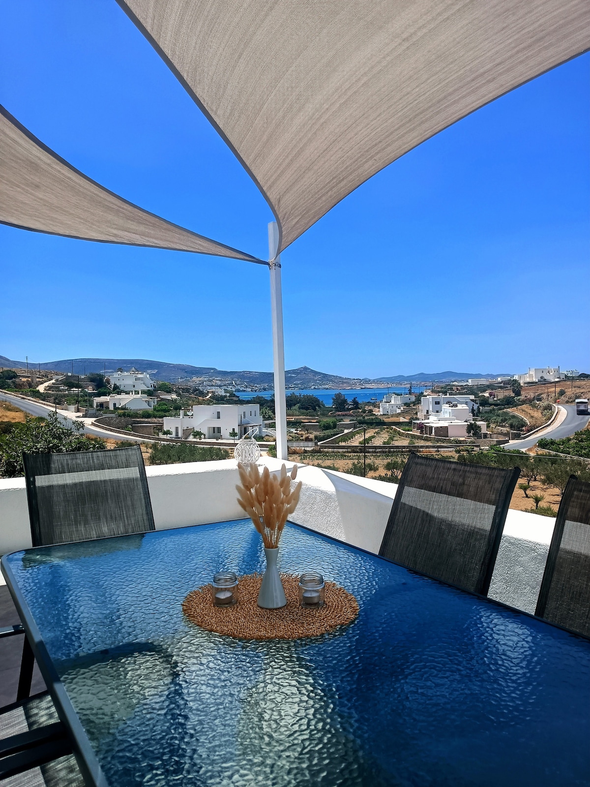 An outdoor dining area is displayed under a shaded canopy, featuring a glass-topped table surrounded by six chairs. A vase with decorative elements rests at the center, while a distant view of the sea and surrounding landscape is visible beyond.