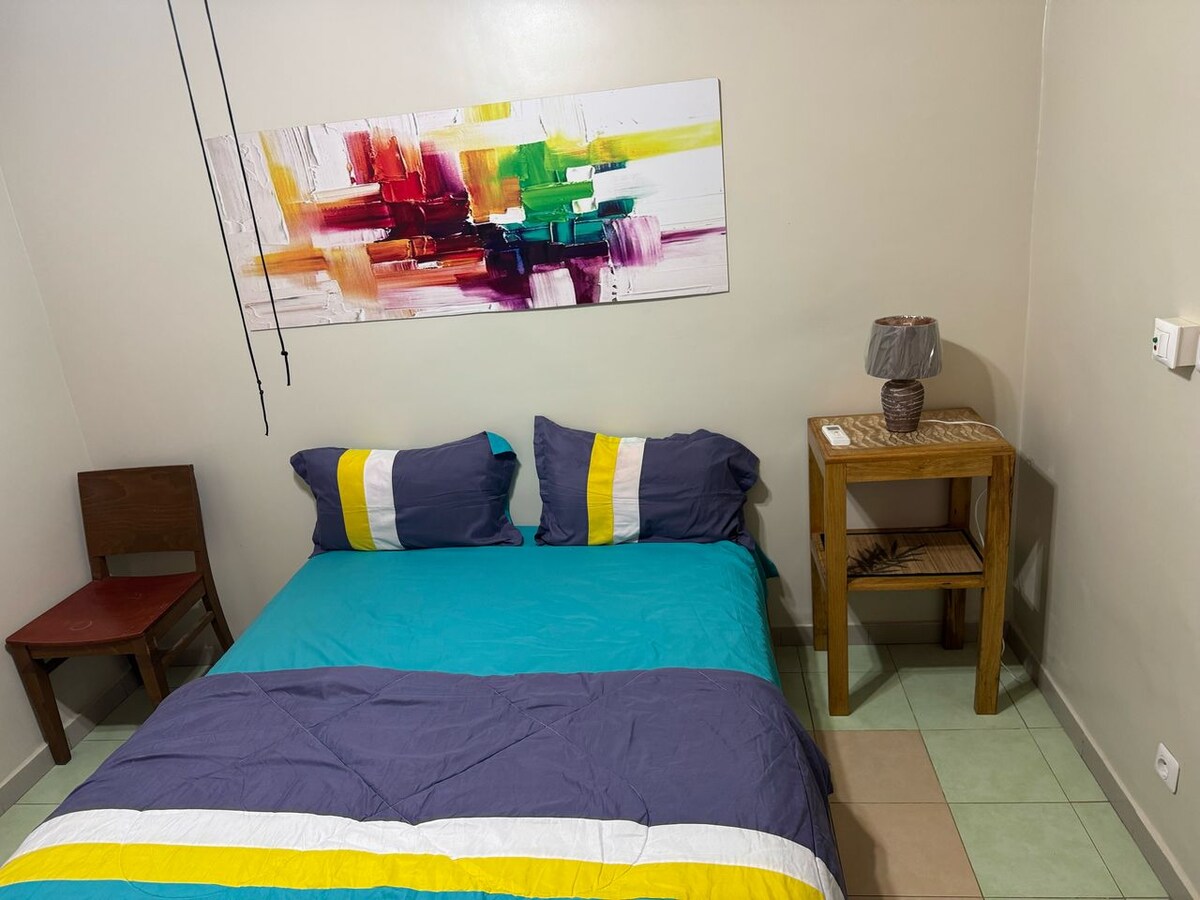 A comfortable sleeping area is presented, featuring a spacious bed with colorful bedding in shades of blue and yellow. A vibrant piece of abstract art hangs above the bed, while a small wooden table and a chair are positioned nearby. Soft lighting is provided by a lamp on the table.