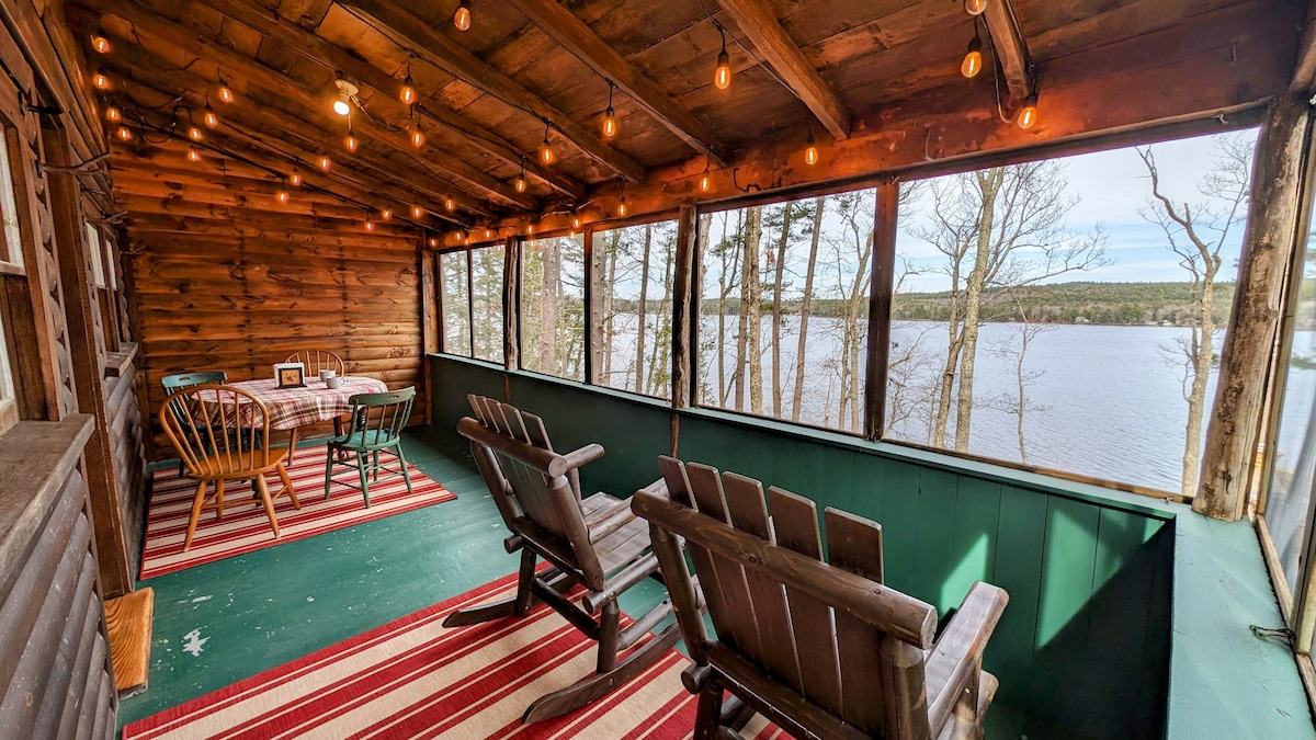 A screened-in porch offers views of the lake, featuring wooden chairs arranged for relaxation. A dining table for four is positioned next to the window, surrounded by natural wood walls and illuminated by string lights. A striped rug adds comfort to the space.