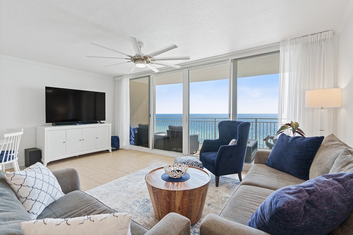 The living area is designed with large glass doors providing unobstructed views of the Gulf. A light-colored sofa, armchairs, and a wooden coffee table are arranged on a neutral-toned rug, complementing the serene atmosphere. A television is positioned on the opposite wall.