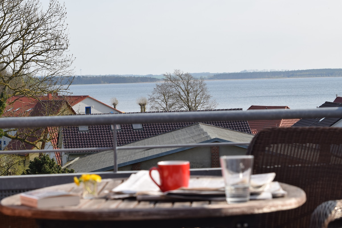 A balcony view captures a serene scene of the water and distant landscape. A small table is set with a red mug and a glass, accompanied by a flower vase. Nearby rooftops and trees frame the image, creating a sense of tranquility.