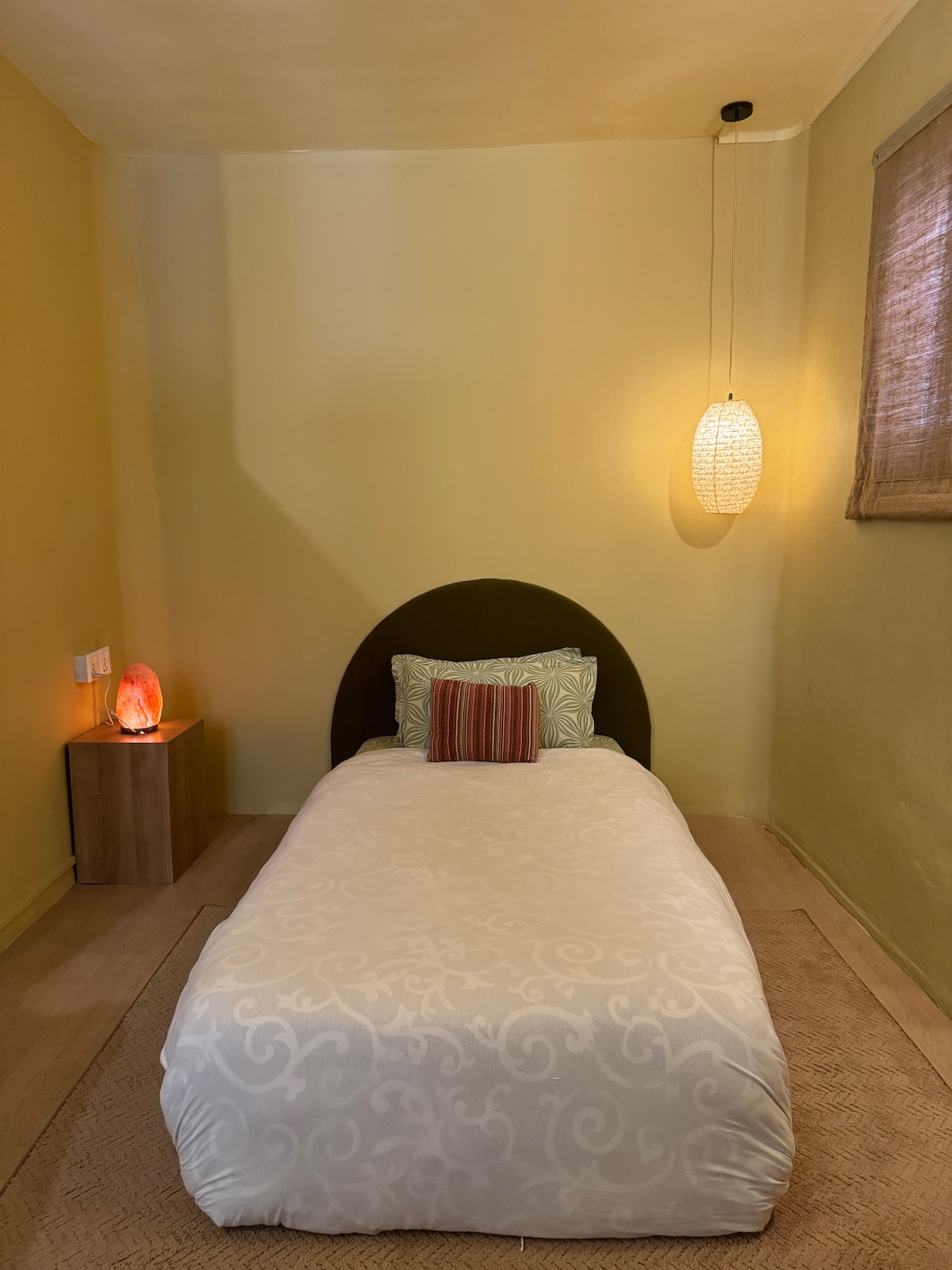 A single bedroom features a spacious bed with a light-colored bedspread and a decorative pillow. A bedside table with a soft glow lamp is positioned nearby. Natural light enters through a window adorned with a simple shade, contributing to the room's calm ambiance.