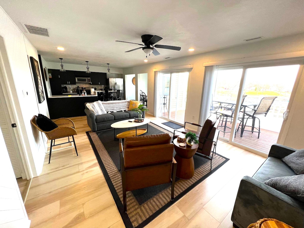 A modern living area showcases comfortable seating arrangements, including a brown armchair and two gray sofas. A central coffee table is positioned on a patterned area rug. Large sliding doors provide access to an outdoor deck, allowing natural light to fill the space.