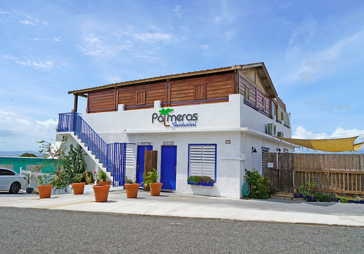 The exterior of Palmeras Beach Apartments features a two-story building with a white facade and wooden accents. Colorful planters adorn the entrance, while a blue staircase leads to the upper level. Vibrant greenery surrounds the property, enhancing its inviting appearance.