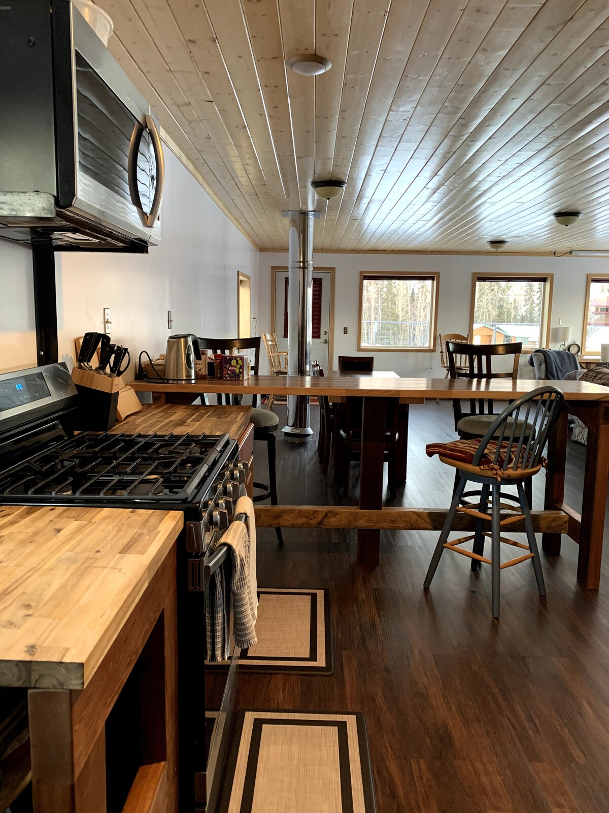 The kitchen space features a wooden countertop with a gas stove and ample cabinetry. A cozy dining area is visible with two dark wooden tables and several chairs arranged around. Natural light enters through large windows, enhancing the inviting atmosphere.