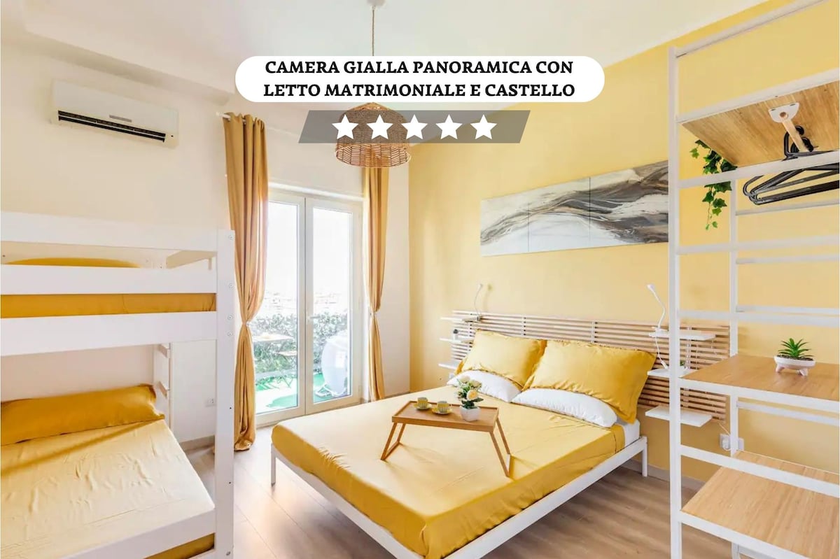 A bright yellow room features a spacious double bed alongside a bunk bed. Large windows allow natural light to fill the space, while a decorative light fixture hangs above. Green plants add a touch of freshness, creating a welcoming atmosphere.