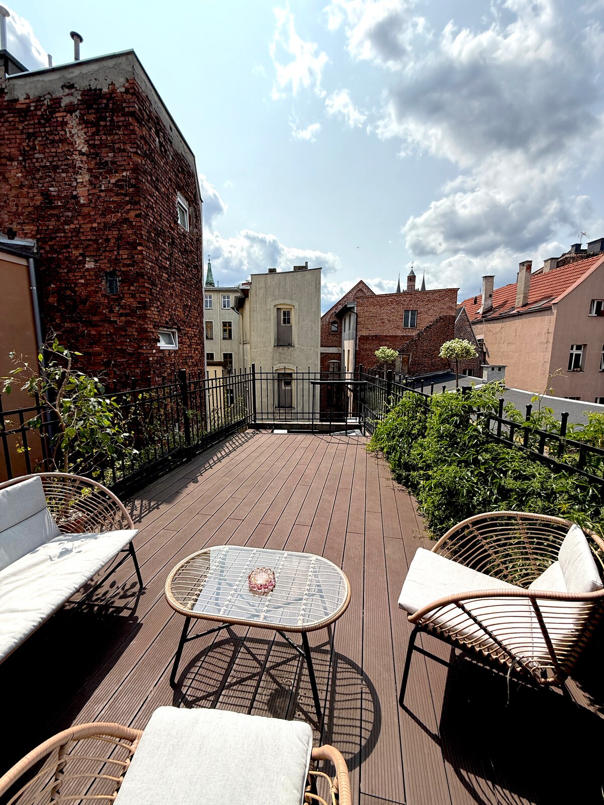 A spacious roof terrace is presented, featuring comfortable seating arrangements with two chairs and a small table. Lush greenery surrounds the area, and the view includes charming neighboring buildings under a partly cloudy sky.