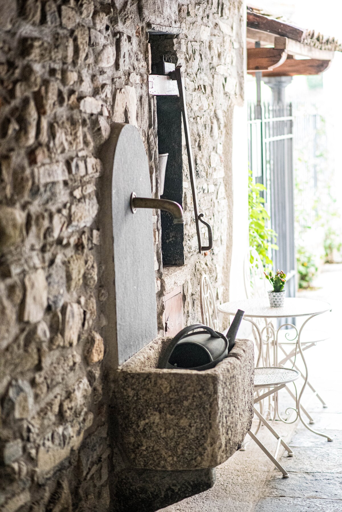 A rustic stone wall features a traditional water trough, made of granite, with a metal spout positioned above it. Nearby, a small round table with white wrought iron chairs provides a cozy seating area. Natural light filters in, enhancing the serene ambiance.